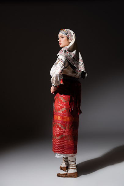 full length of young ukrainian woman in traditional dress with ornament and headwear posing on black - Foto, afbeelding