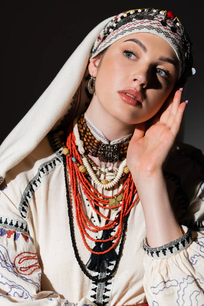 portrait of ukrainian woman in traditional shirt with red ornament and headwear looking away isolated on black - Foto, Bild