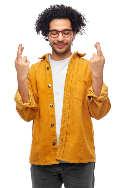 gesture, luck and people concept - smiling young man with crossed fingers in yellow jacket over white background - 写真・画像