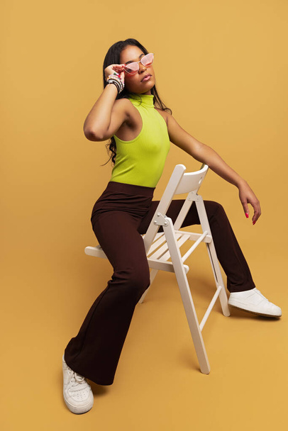 Full body of young Latin American female in stylish clothes sitting on chair and adjusting sunglasses against beige background - Zdjęcie, obraz