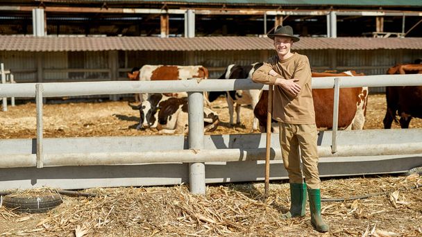 Smiling male farmer with rake standing near paddock with herd of milk cows on farm. Young caucasian guy looking at camera. Modern hipster countryside lifestyle. Agriculture. Sunny day - Photo, image