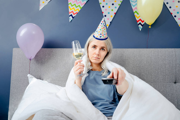 Sad woman in pajamas, party cap drinks champagne and watches TV, wrapped in a blanket while sitting on a bed in a decorated bedroom. Celebrates a birthday alone. Boring home party. Selective focus. - Foto, Imagen