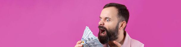 Portret zadowolonego młodego biznesmena z brodą ubranego w różową kurtkę, pokazującego banknoty dolarowe na czerwonym tle studia. Smak, zapach pieniędzy. - Zdjęcie, obraz