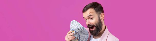 Ritratto di un giovane uomo d'affari soddisfatto con la barba vestita con una giacca rosa che ci mostra banconote in dollari su uno sfondo rosso da studio. Gusto, odore di soldi. - Foto, immagini