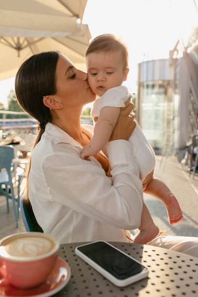 Young tanned woman with long dark hair wearing white shirt holding her baby boy and kissing on summer terrace during walking. Caucasian woman drinking coffee on terrace with baby. - Foto, Bild