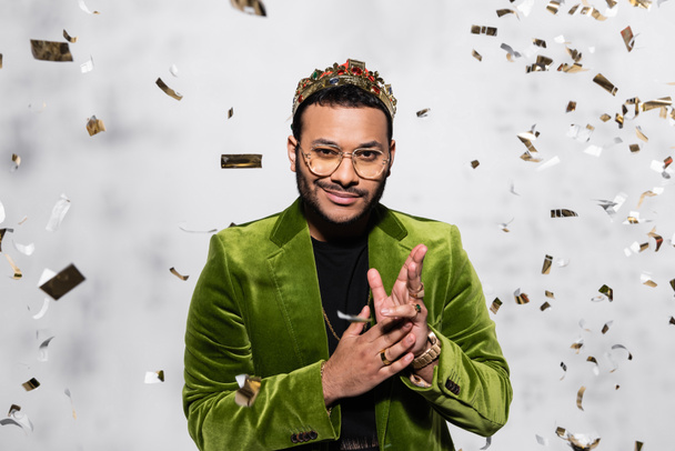 pleased indian hip hop performer in green velvet blazer and crown near falling confetti on grey - Foto, afbeelding