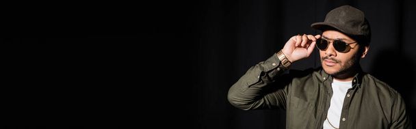 eastern hip hop performer in cap adjusting sunglasses on black, banner - Valokuva, kuva