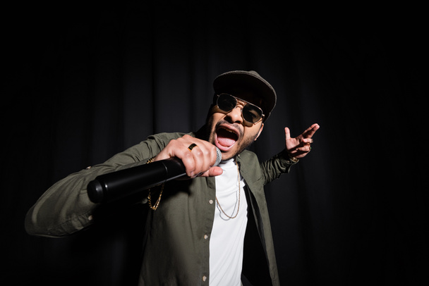 indian hip hop performer in sunglasses and cap singing in microphone on black - Fotografie, Obrázek