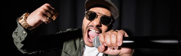 Індіанський хіп-хоп виконавець у сонцезахисних окулярах і кашкет співають гучно у мікрофоні на чорному, банер - Фото, зображення