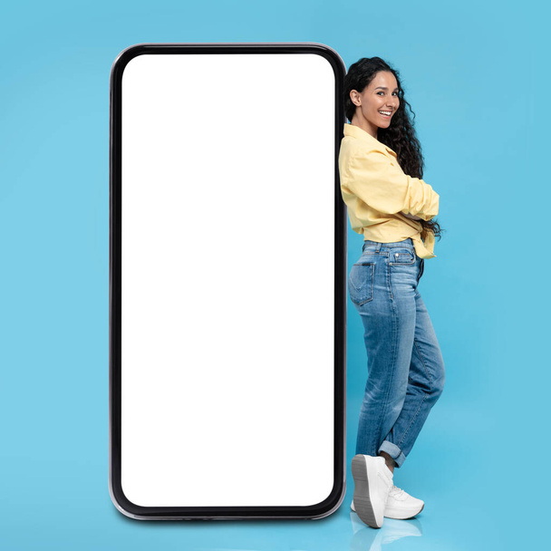 Счастливая ближневосточная женщина опирается на огромное рекламное приложение Blank Screen или мобильное предложение, стоящее на голубом студийном фоне. Макет, квадратный шот - Фото, изображение