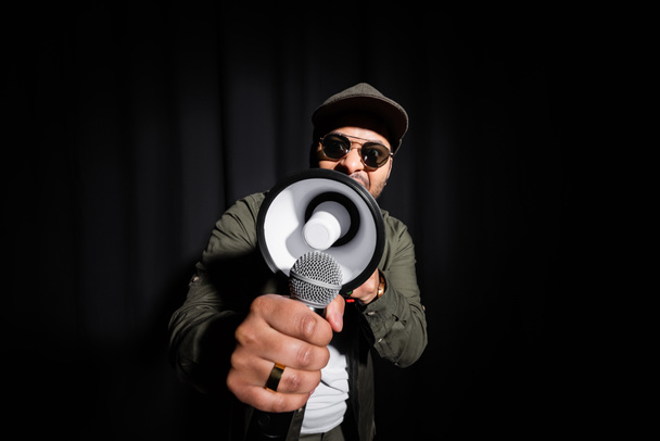 middle east hip hop performer in sunglasses and cap singing in microphone and loudspeaker on black - Fotoğraf, Görsel