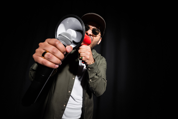 middle east hip hop singer in sunglasses and cap singing in microphone and loudspeaker on black - Foto, imagen