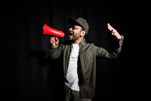 emotional middle east hip hop singer in sunglasses talking in loudspeaker on black - Zdjęcie, obraz