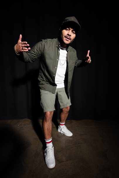 full length of stylish indian hip hop singer in cap gesturing while standing on black  - 写真・画像