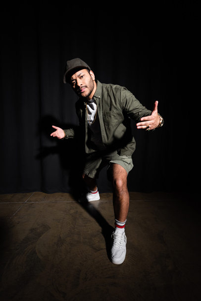 full length of stylish eastern hip hop singer in cap gesturing while standing on black  - Fotoğraf, Görsel