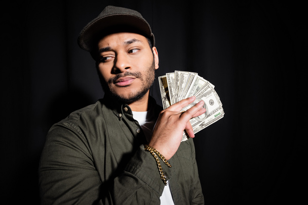 elegante cantante indio de hip hop con gorra sosteniendo billetes de dólar aislados en negro  - Foto, imagen