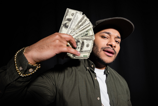 bearded indian hip hop singer in cap holding dollar banknotes isolated on black  - Valokuva, kuva