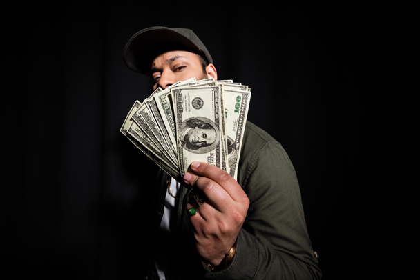 bearded indian hip hop performer in cap covering face with dollar banknotes on black  - Photo, Image