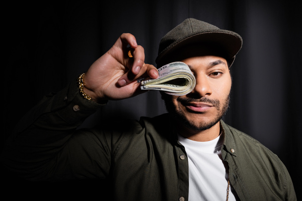 rich indian hip hop performer in cap covering eye with bundle of dollar banknotes on black  - Fotoğraf, Görsel