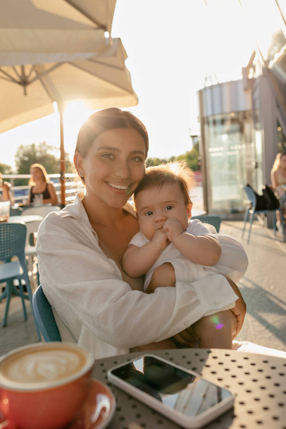 Outdoor portrait of happy young mom with baby boy posing with smile on summer terrace in sunlight. Charming woman relaxing in the city with her son on sunrise - Foto, Bild