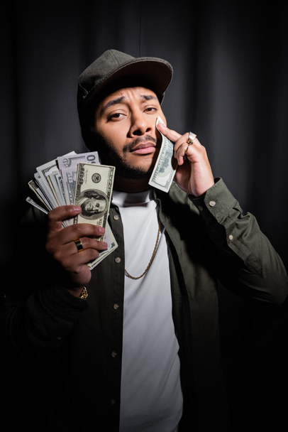 sad indian hip hop performer in cap wiping tear with dollar banknote on black  - Fotó, kép