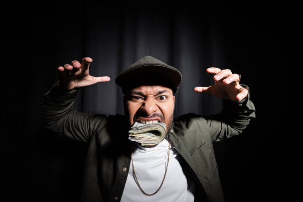 rich indian hip hop performer in cap biting bundle of dollar banknotes and showing spooky gesture on black  - Fotografie, Obrázek