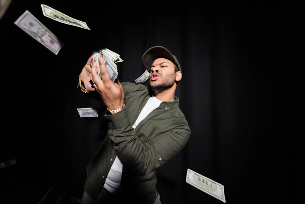 rich indian hip hop performer in cap throwing blurred dollar banknotes on black  - Zdjęcie, obraz
