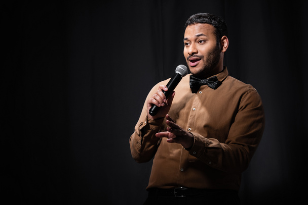 indian comedian in shirt and bow tie gesturing and holding microphone during monologue on black  - Fotografie, Obrázek
