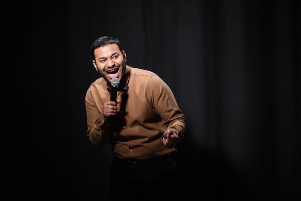 amazed indian stand up comedian telling jokes into microphone on dark stage on black  - Fotografie, Obrázek