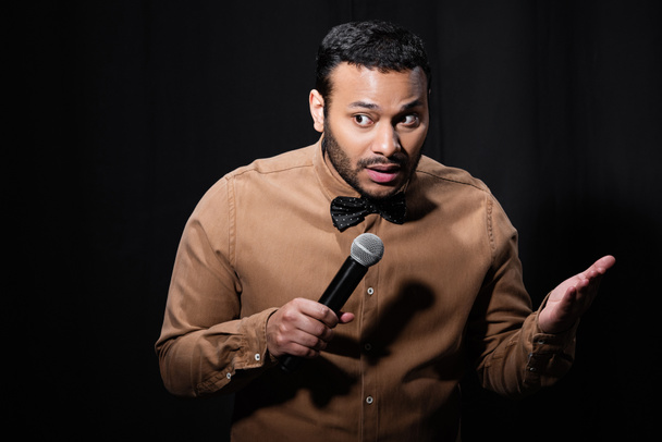 indian stand up comedian gesturing while telling jokes into microphone on dark stage isolated on black  - Fotoğraf, Görsel