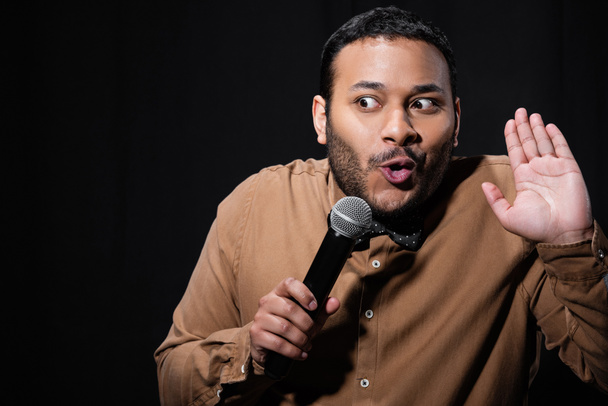 emotional indian stand up comedian showing stop gesture while telling jokes into microphone isolated on black  - Foto, Imagen