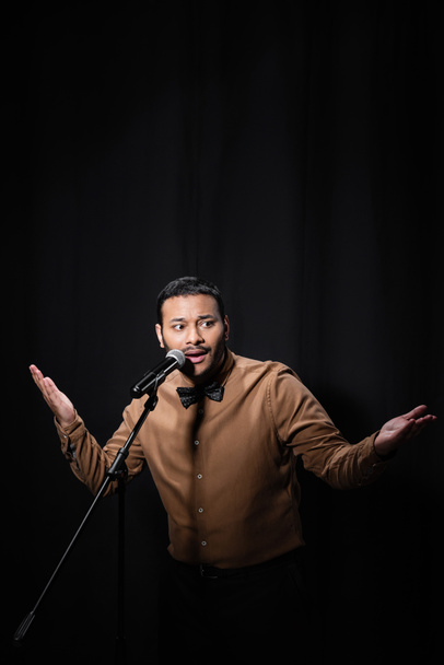 indian stand up comedian gesturing while telling jokes into microphone on stand isolated on black  - Φωτογραφία, εικόνα
