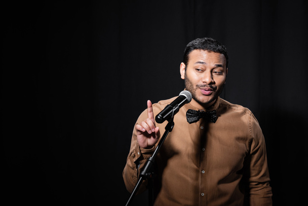 indian stand up comedian pointing with finger while telling jokes into microphone on stand on black background - Photo, Image