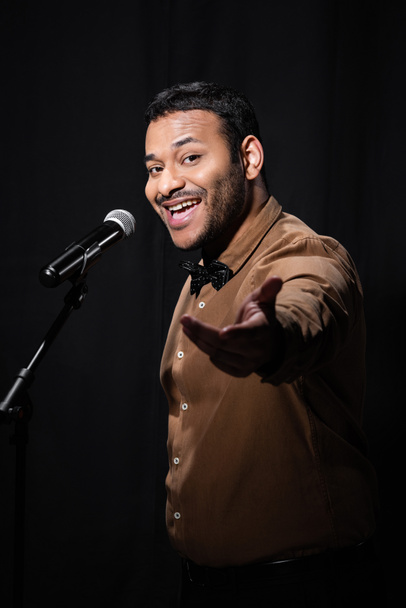 amazed indian stand up comedian telling jokes into microphone on stand isolated on black  - Foto, Imagen