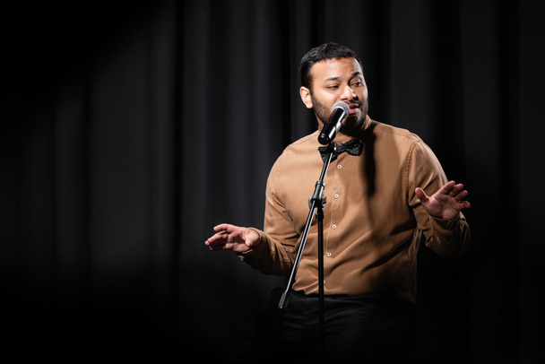 indian comedian gesturing and performing stand up comedy while telling jokes in microphone on black  - Valokuva, kuva