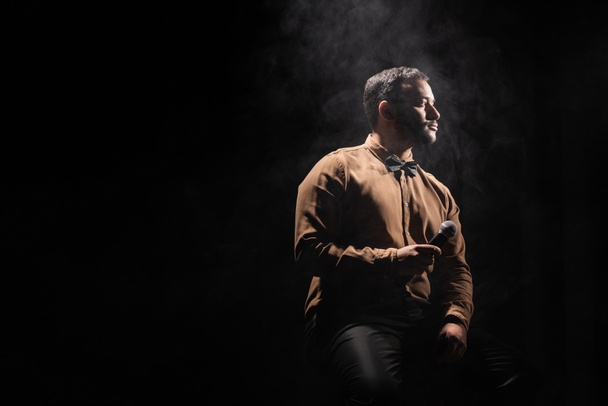indian comedian holding microphone and looking away on black with smoke - Foto, afbeelding