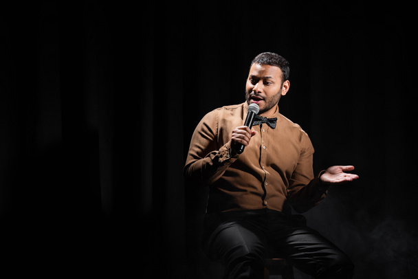 indian comedian sitting and performing stand up comedy into microphone on black  - Fotoğraf, Görsel