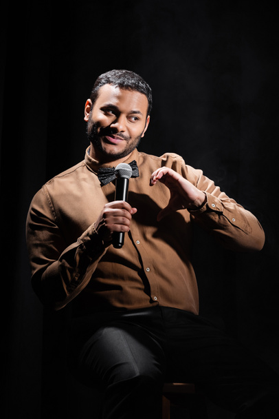 smiling indian comedian sitting and performing stand up comedy on black  - Foto, immagini