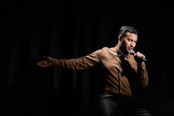 eastern comedian sitting and performing stand up comedy into microphone on black  - Foto, Imagen