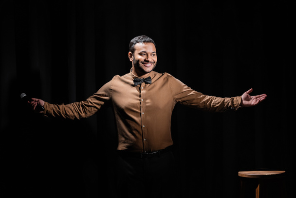 happy middle east comedian gesturing and holding microphone on black  - 写真・画像