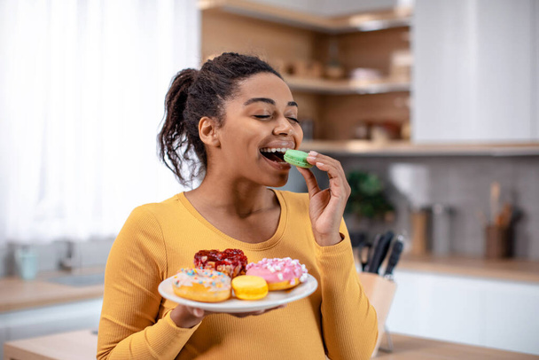 Bin yıllık hamile Afro-Amerikan kadının elinde bir tabak şeker tutmasına ve minimalist mutfak içinde kurabiye yemesine sevindim. Tatlıya düşkün ve abur cubur düşkünü, bebek için covid-19 virüsü bekleniyor. - Fotoğraf, Görsel