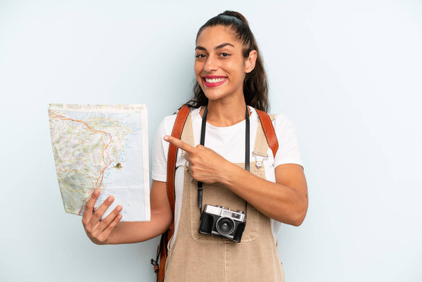 hispanic woman smiling cheerfully, feeling happy and pointing to the side. tourist with a map - Foto, afbeelding