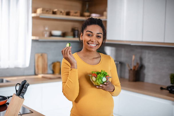 Glad hungry pretty millennial pregnant african american woman with belly eating salad from vegetables in modern kitchen interior. Proper nutrition, diet, health care, vegetarian for unborn child - Foto, imagen