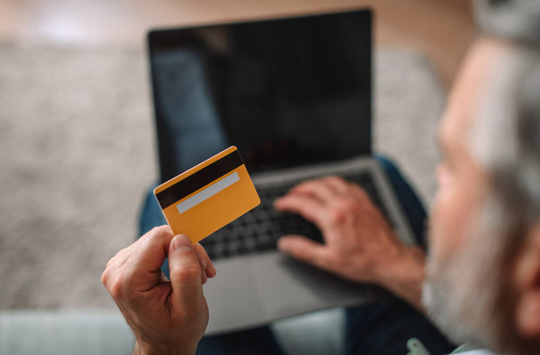 European senior male with beard looks at laptop with empty screen, shows credit card in living room interior, close up. Offer, new website, online shopping, sale and technology due covid-19 quarantine - Foto, Imagen