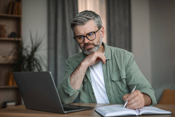 Serious caucasian male with beard in glasses looks at laptop have video call, study in living room interior. Pensioner work at home with device, meeting remotely and business due covid-19 pandemic - Foto, imagen