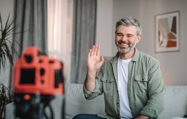 Smiling caucasian mature male with beard waving hand at camera, shooting video in living room interior. Modern work remote, new tech, hobbies and new normal, blogging at home due covid-19 quarantine - Fotografie, Obrázek