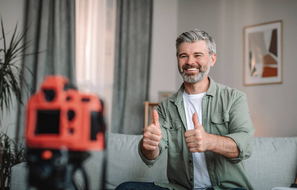 Glad happy caucasian old male blogger with beard showing thumbs up and shooting video in living room interior. New technology, hobbies and new normal, blog and social distance due covid-19 pandemic - Photo, Image