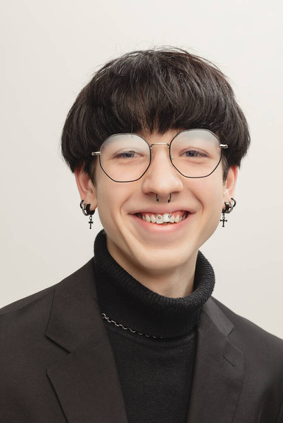 Portrait of young stylish man with piercing, in glasses posing, cheerfully smiling isolated over grey studio background. Concept of modern fashion, art photography, style, queer, uniqueness, ad - Photo, Image