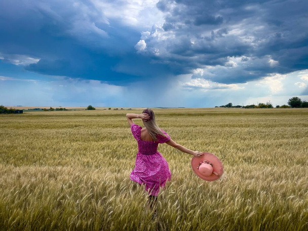 Женщина в розовом платье и шляпе в поле пшеницы в облачный и бурный летний день - Фото, изображение