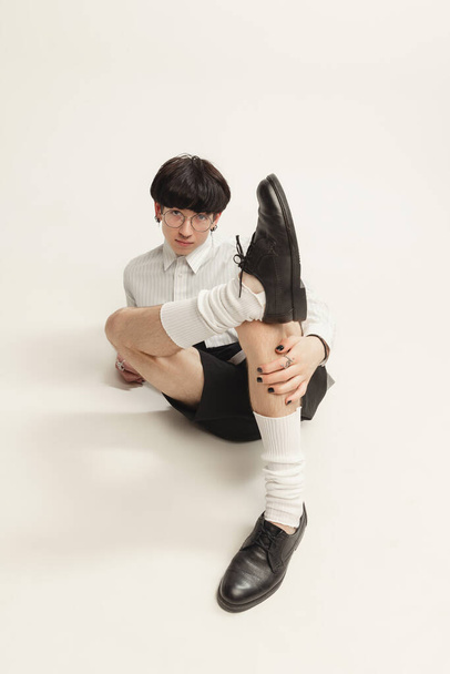 Porträt eines jungen Mannes im schwarz-weißen Outfit mit Brille, der auf dem Boden sitzt und isoliert vor grauem Studiohintergrund posiert. Konzept der modernen Mode, Kunstfotografie, Stil, Queer, Einzigartigkeit, Werbung - Foto, Bild